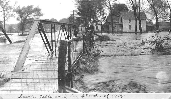 Above: Excerpt of a photo of the 1915 Flood of Table Rock’s Lower Town. The view is from the west side of the Taylor Branch bridge. Note the daring young man on the bridge railing above the standing wave of flood waters hitting the bridge. The house in the foreground is gone, replaced by a small house built by Albert &amp;amp; Bernice Hillers. Donnie Kalina lives there now. The author lives in the next house after that, and she sometimes winces during heavy rains as she thinks of this picture.