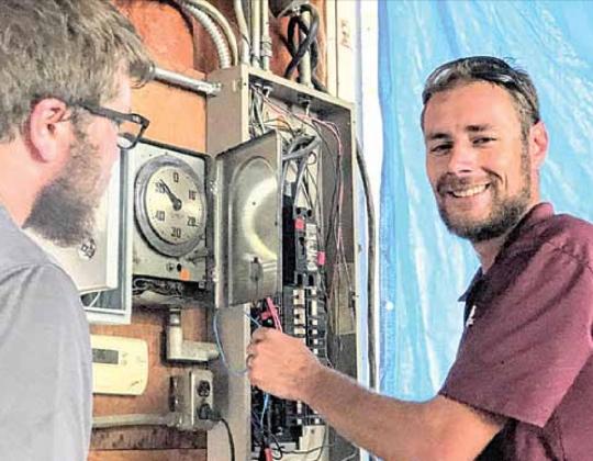 Daniel Schell of Haug Communications at work, co-worker Jonathon Johnson at left. The clock face is part of the present system, which is supplemented by a new control installed to its left, breaker box to the right. Sharla Sitzman Photo