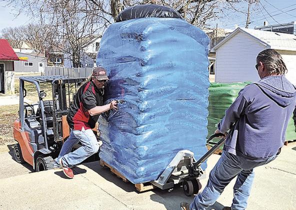 R.J. Hickey, assistant manager of the Pawnee City True Value store stands on the sidewalk in front of the store next to stacks of products and employees Nick Klein (left) and Gary Cumley (right) strain to move a load of mulch which is stored in the back of the buiding,