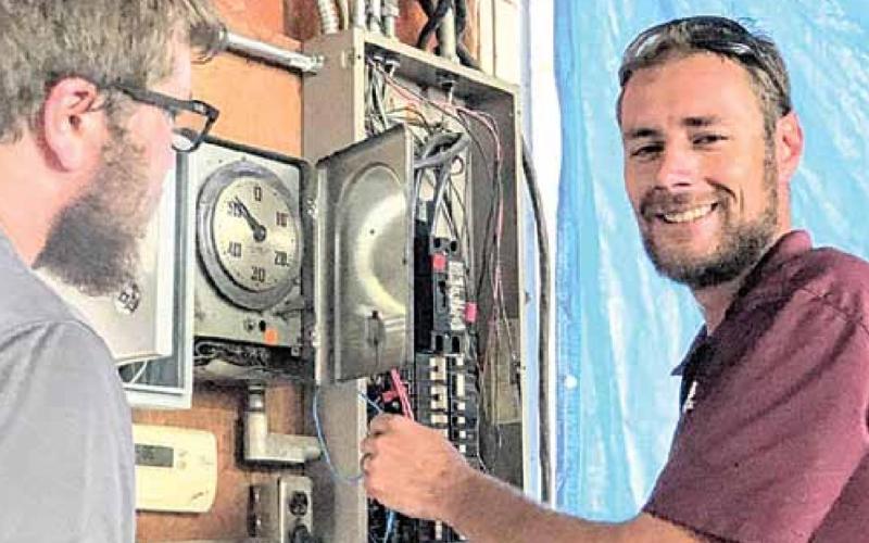 Daniel Schell of Haug Communications at work, co-worker Jonathon Johnson at left. The clock face is part of the present system, which is supplemented by a new control installed to its left, breaker box to the right. Sharla Sitzman Photo