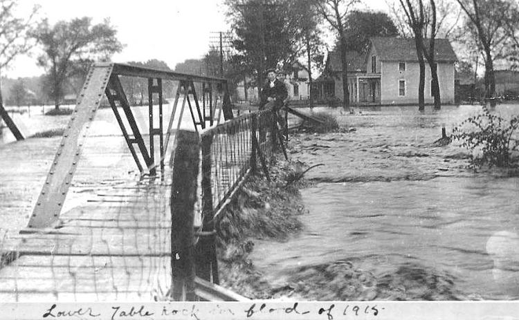 Above: Excerpt of a photo of the 1915 Flood of Table Rock’s Lower Town. The view is from the west side of the Taylor Branch bridge. Note the daring young man on the bridge railing above the standing wave of flood waters hitting the bridge. The house in the foreground is gone, replaced by a small house built by Albert &amp; Bernice Hillers. Donnie Kalina lives there now. The author lives in the next house after that, and she sometimes winces during heavy rains as she thinks of this picture.