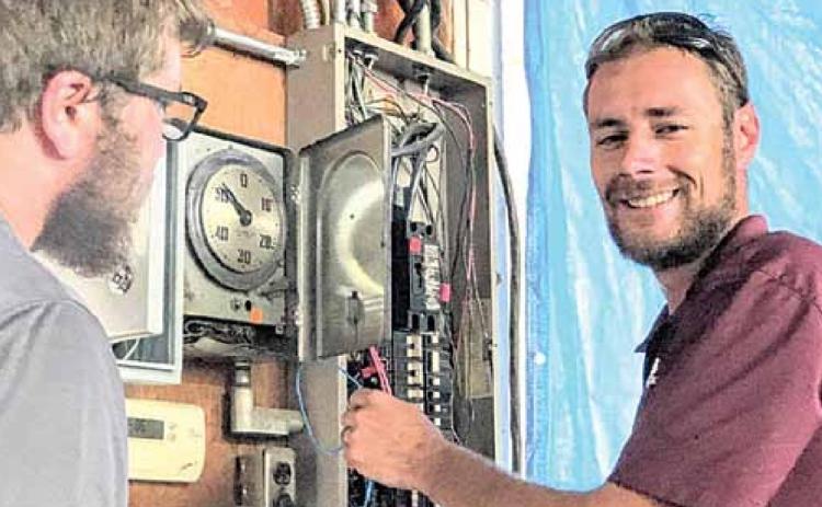 Daniel Schell of Haug Communications at work, co-worker Jonathon Johnson at left. The clock face is part of the present system, which is supplemented by a new control installed to its left, breaker box to the right. Sharla Sitzman Photo